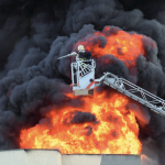 masive fire with black smoke and huge flames and a crane above the fire with a firefighter trying to put it out with a hose