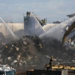 image of firefighters on cranes, using a fire hose to put out a fire in a junk yard