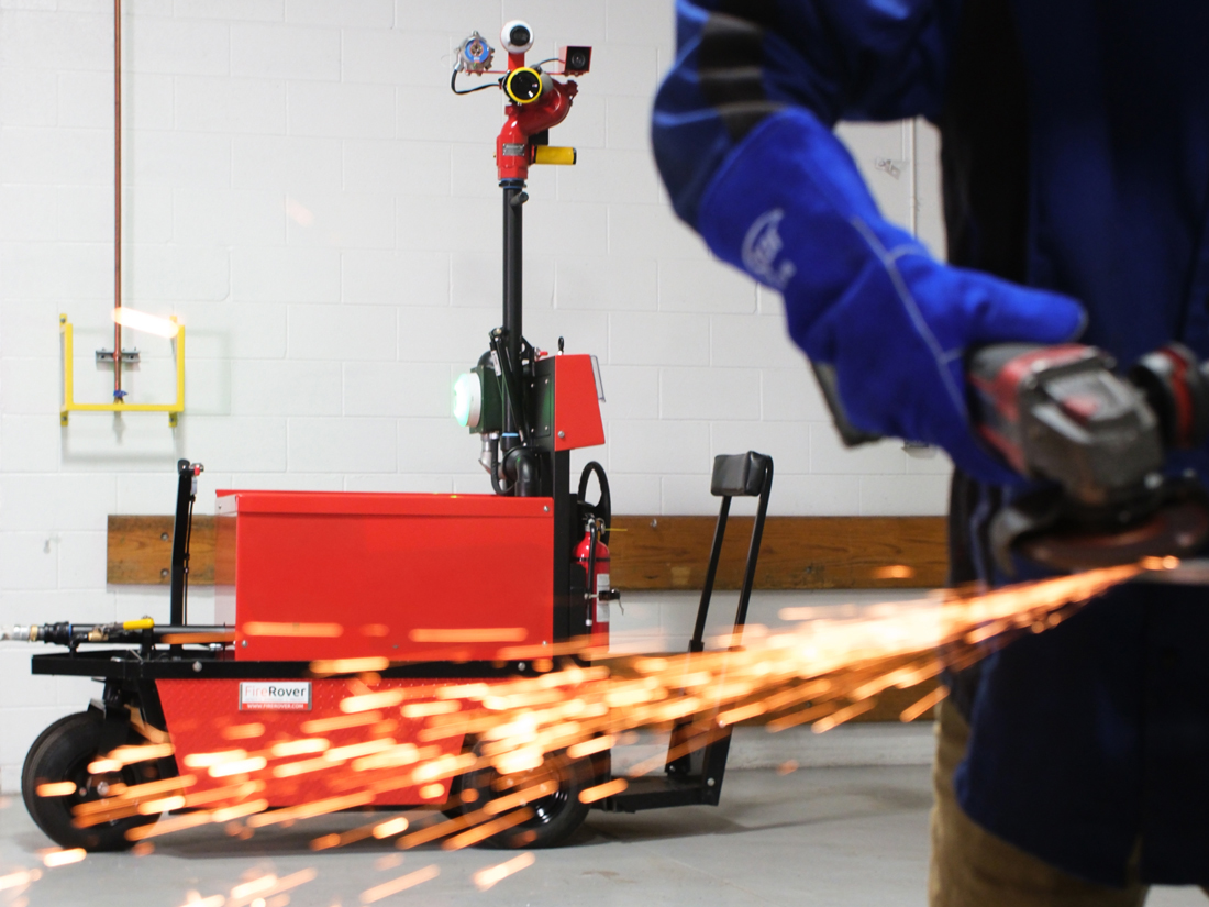 A Fire Rover mobile suppression unit positioned on a concrete floor monitors a worker in blue welding gloves using an angle grinder, with a dramatic stream of orange sparks flying toward the unit during a hot work safety demonstration.