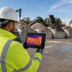 Construction worker is monitoring a thermal imaging video feed on his tablet, with an organized construction site in the background.