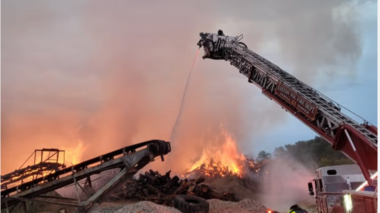 Firefighters use an aerial ladder truck to spray water on a large fire at a waste or recycling facility as heavy smoke rises.